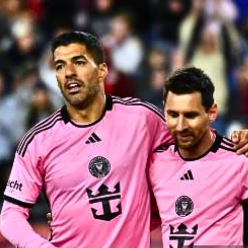Lionel Messi and Luis Suárez standing side by side wearing Inter Miami’s iconic pink jerseys during a Major League Soccer match in the United States, smiling and looking ready to play, with a packed stadium and cheering fans in the background.