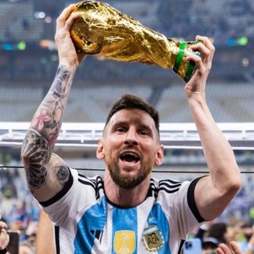 Lionel Messi lifting the FIFA World Cup trophy high above his head after Argentina’s dramatic victory over France in the 2022 World Cup final in Qatar, surrounded by cheering teammates like Ángel Di María, Julián Álvarez, and Emiliano Martínez. Confetti rains down inside the packed Lusail Stadium as Messi, wearing Argentina’s iconic blue and white striped jersey, smiles with pure joy and relief, finally achieving his lifelong dream and cementing his place as one of the greatest footballers in history.